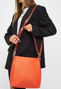 Oranje synthetische handtas met een gladde textuur, bruine faux leren schouderriem, gouden hardwareaccenten en logo aan de voorkant.