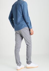 Man wearing a blue button-up shirt, gray slim-fit pants, and white sneakers, standing against a plain light background.