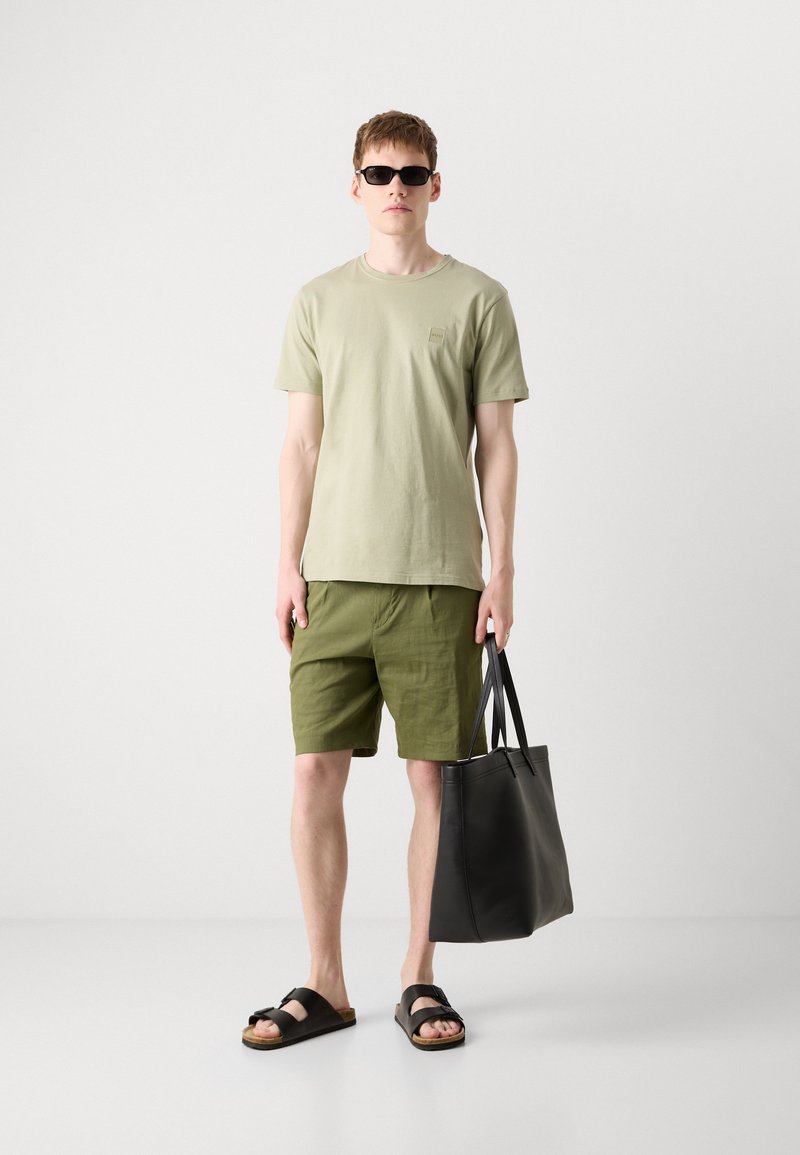 T-shirt vert clair à manches courtes, shorts vert olive, sac fourre-tout noir et sandales noires ; design simple avec des accents minimaux.