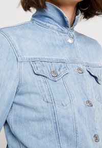 Light blue denim jacket with a high collar, silver snap buttons, and dual chest pockets. Features a textured finish and tailored seams.