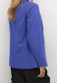 Blazer su misura in tessuto blu brillante con una texture liscia; presenta una ventola singola, rever a punta e maniche lunghe.