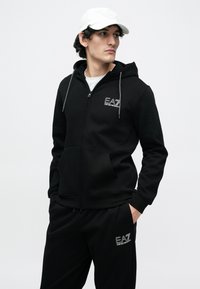 Νεαρός άνδρας φοράει μαύρη ζακέτα με φερμουάρ EA7 Emporio Armani και παντελόνια, λευκό καπέλο, κοιτάζει στο πλάι, με τα χέρια στις τσέπες μπροστά σε απλό φόντο.