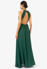 Abito verde lunghi a terra con schiena drappeggiata, spalline a halter plissettate e una silhouette fluida. Tessuto morbido con una texture liscia.