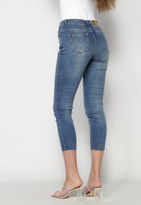 Jeans de mezclilla en un color azul desgastado, con un diseño ajustado, de largo hasta el tobillo y con dobladillo sin terminar. Presenta dos bolsillos traseros con costuras decorativas.