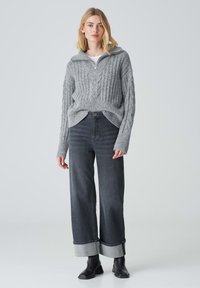 Maglione grigio a maglia con zip parziale, caratterizzato da motivi a treccia; abbinato a jeans scuri larghi con polsini a contrasto e stivaletti neri.