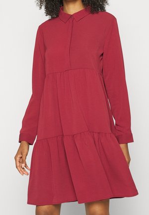 Mujer que lleva un vestido largo hasta la rodilla de manga larga, de color rojo oscuro con volantes, con cuello puntiagudo y botones delanteros ocultos.