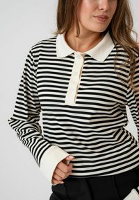 Polo majica dugih rukava crno-bijelih pruga s krem okovratnikom, s patent zatvaračem i rebrastom teksturom. Rukavi su krem boje.