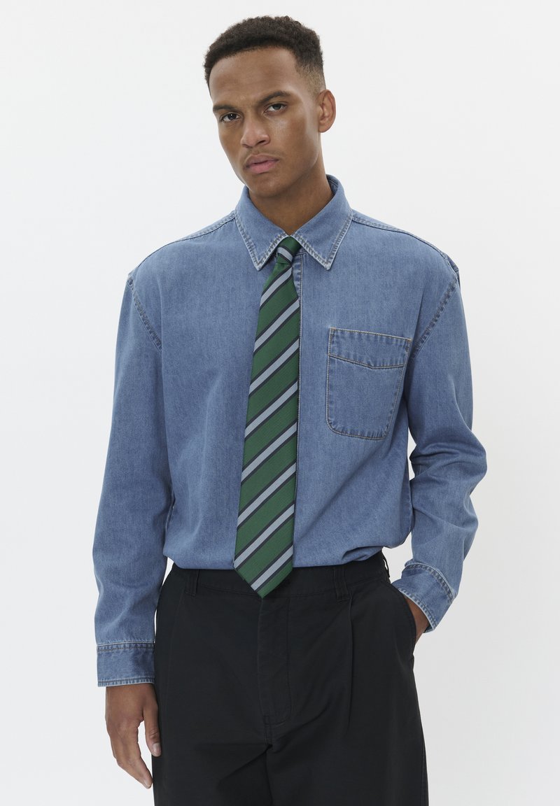 Denim särg terava kraega, pikkade varrukatega ja rinna-taskuga. Kombineeritud rohelise ja halli triibulise lipsuga. Mustad püksid. Minimalistlik disain.