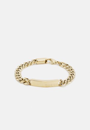 Guldfarvet kædearmbånd med en flad ID-plade indgraveret med "GUESS" og en sikker lukning. Designet er i metal med en blank overflade.