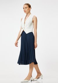 Witte mouwloze blazer met een schuilkraag, gecombineerd met een knielange marineblauwe geplooide rok en witte puntige hakken met strikaccenten.