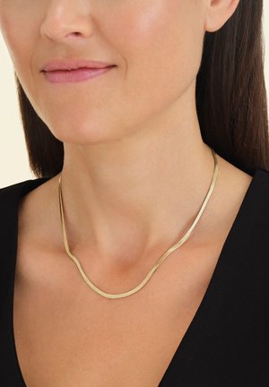 Gouden metalen ketting met een glad, golvend ontwerp, rustend op een blote sleutelbeen. Achtergrond is neutraal, waardoor de sieraden opvallen.