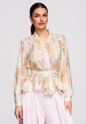 Femme portant un chemisier cache-cœur à manches longues à motifs floraux avec une ceinture et un pantalon plissé taille haute rose clair, debout devant un fond uni.