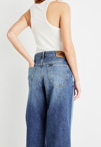 Žena v bílém žebrovaném topu a modrých džínech Lee s vysokým pasem a širokými nohavicemi, stojící s jednou rukou v zadní kapse, otočená zády.