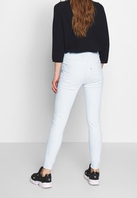 Nainen, jolla on pitkät hiukset, pukeutuneena mustaan lyhyeen neuleeseen, vaaleansinisiin skinny-farkkuihin ja mustiin Adidas-tennareihin, seisoo tasaisessa valkoisessa taustassa.