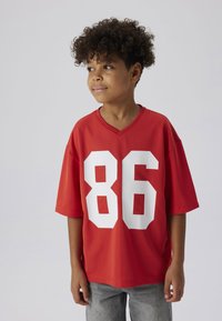 Rode korte mouwen shirt met een V-hals en groot wit nummer "86" op de voorkant. De stof lijkt ademend en heeft een textuur. Draagt met grijze jeans.