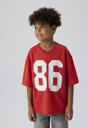 Rode korte mouwen shirt met een V-hals en groot wit nummer "86" op de voorkant. De stof lijkt ademend en heeft een textuur. Draagt met grijze jeans.