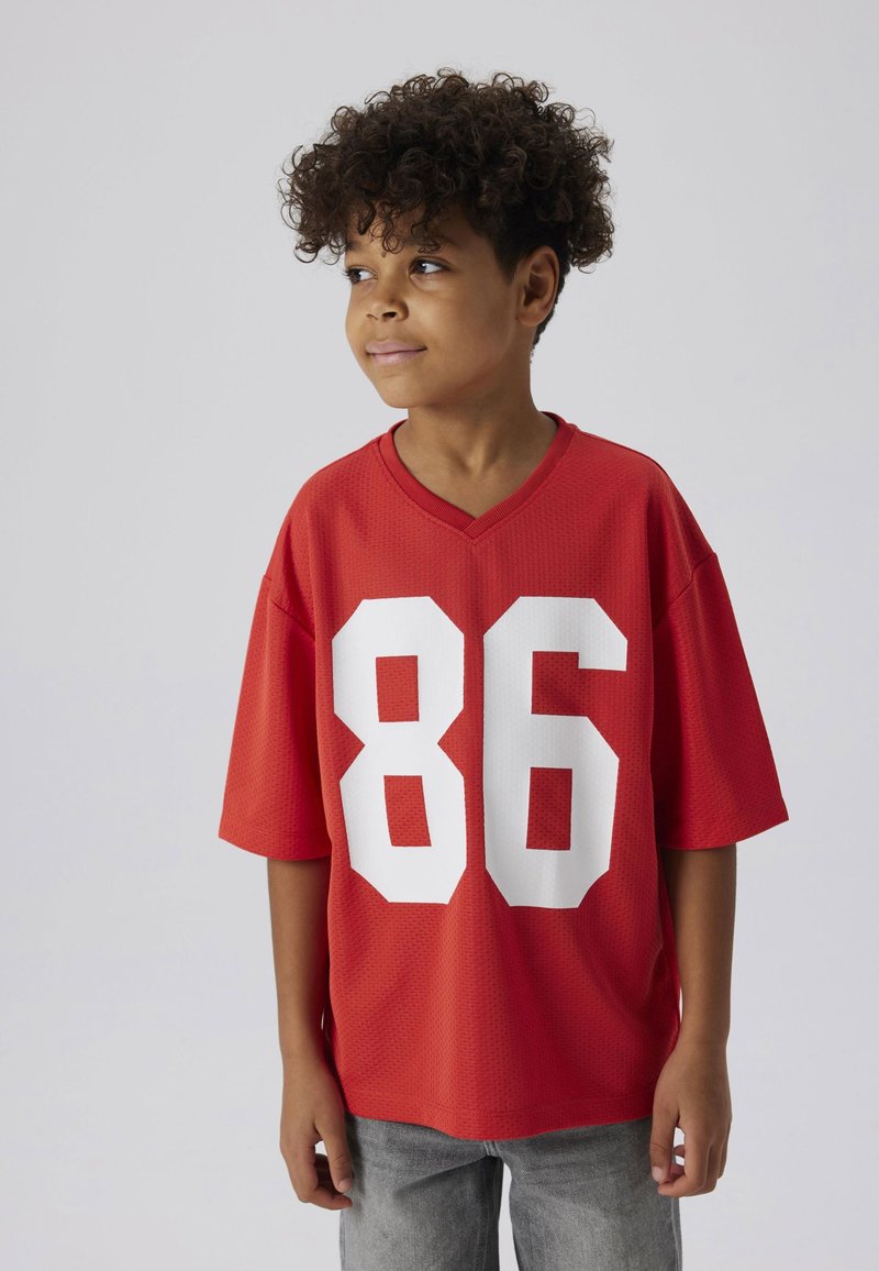 Rode korte mouwen shirt met een V-hals en groot wit nummer "86" op de voorkant. De stof lijkt ademend en heeft een textuur. Draagt met grijze jeans.