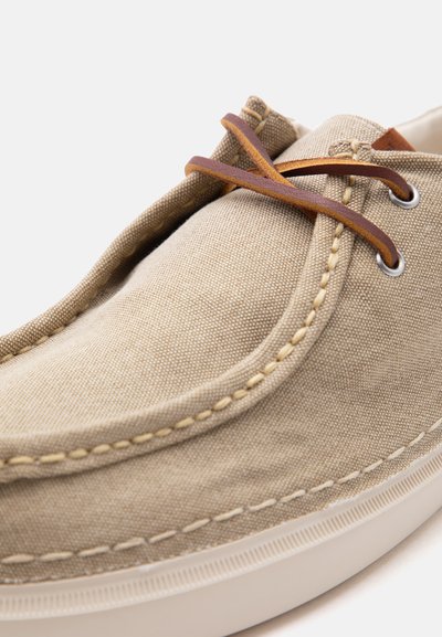 Μπεζ υφασμάτινο καθημερινό παπούτσι με καφέ δερμάτινα κορδόνια, ορατές ραφές, μεταλλικά οφρύδια και λευκή ελαστική σόλα, εμφανιζόμενο σε κοντινή λήψη.