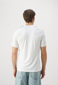 Camiseta atlética blanca con cuello redondo y mangas cortas. Presenta detalles en negro y rojo en los hombros. Tela suave y elástica.