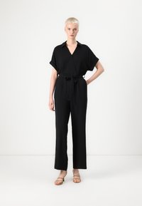 Svart jumpsuit med korte oppbrettede ermer, V-hals og belte i livet. Laget av glatt stoff med bred benåpning og en definert silhuett.