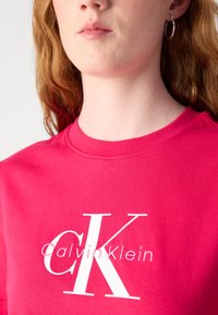 Camiseta de algodón rosa que presenta un gran logo blanco que dice "CK Calvin Klein". El escote es de estilo redondo con una textura suave.