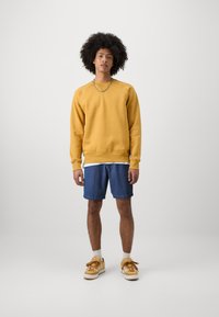 Κίτρινη φόρμα με ραμμένα μανίκια, τζιν σορτς και καφέ αθλητικά παπούτσια. Η φόρμα έχει στρογγυλή λαιμόκοψη, ενώ τα σορτς είναι στο μήκος του γόνατος.