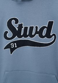 Sweatshirt bleu avec lettrage texturé noir « Stud » et un swoosh noir contenant le chiffre « 91 » au-dessus de la poche avant.