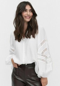 Weiße Bluse mit Puffärmeln, die mit Spitze verziert sind, und Knopfverschluss, kombiniert mit dunkelbraunen, hochgeschnittenen Hosen.