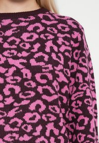 Maglione lavorato a maglia con una base di un viola scuro e una vivace stampa leopardata rosa. Il colletto rotondo e il tessuto texturizzato creano un aspetto accogliente.