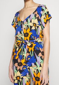 Femme portant une combinaison florale colorée à manches courtes, avec des boutons en bois, un profond col en V et une ceinture nouée à la taille.