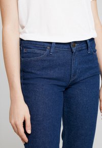 Vaqueros de mezclilla azul oscuro con un corte clásico, que cuentan con cinco bolsillos, costuras sutiles y un cierre de botón metálico en la cintura. Textura suave.