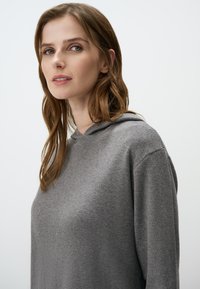 Grijze hoodie met een zachte textuur en een ontspannen pasvorm, met ribbelboorden. Het model heeft golvend haar en een neutrale achtergrond.