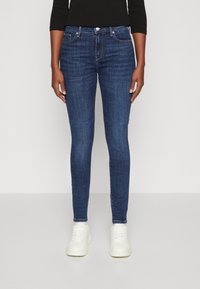 Jeans skinny de denim azul oscuro con un diseño clásico de cinco bolsillos, herrajes plateados y cintura de altura media, combinados con zapatillas blancas.