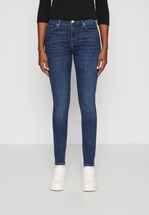 Jeans skinny de denim azul oscuro con un diseño clásico de cinco bolsillos, herrajes plateados y cintura de altura media, combinados con zapatillas blancas.