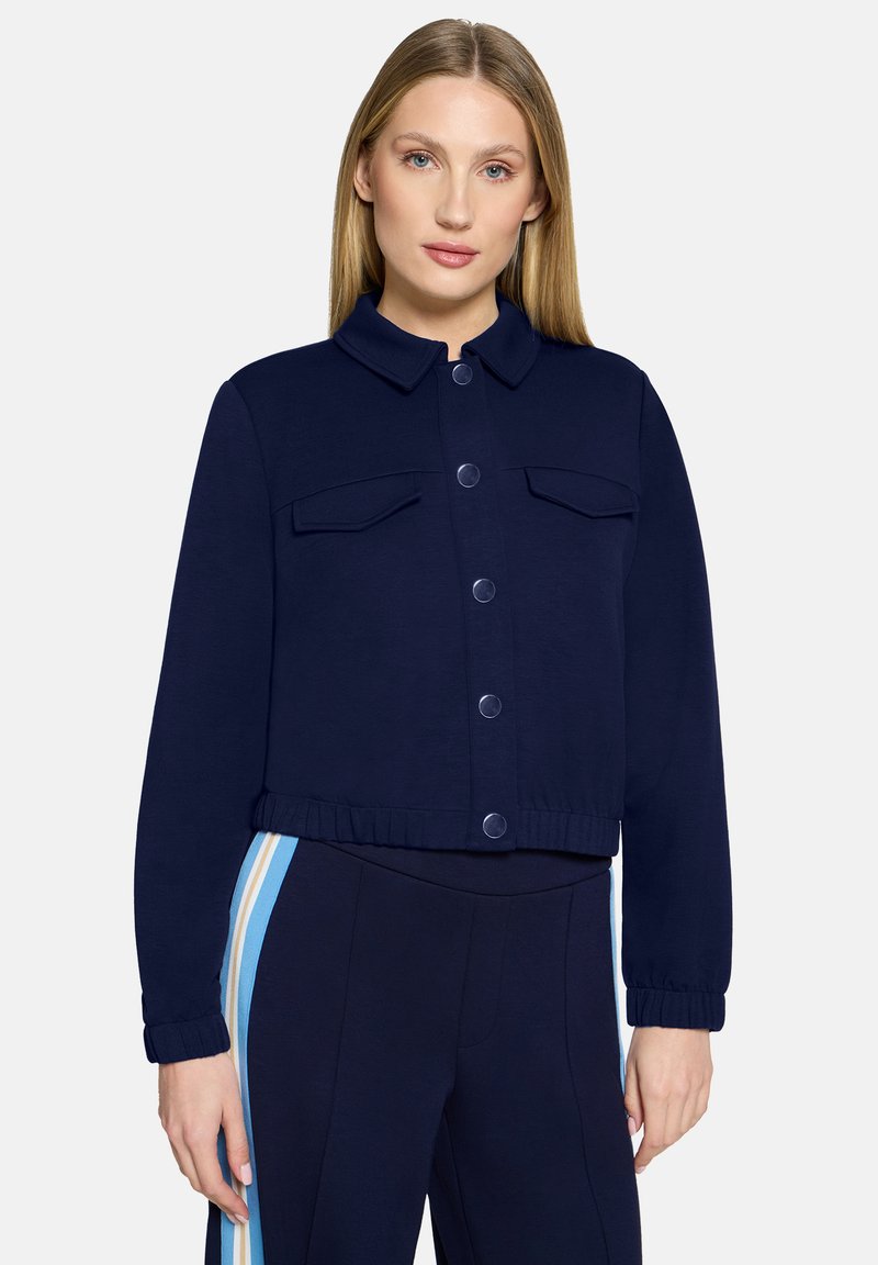 Granatowa kurtka krótkiego kroju z kołnierzykiem, guzikami z przodu i dwoma kieszeniami na piersiach. Wykonana z gładkiej tkaniny z elastycznymi mankietami i dołem.