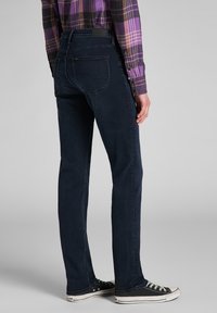 Jean en denim bleu foncé avec une coupe droite, poches arrière et un logo Lee sur la ceinture, accompagnés de baskets en toile noires.