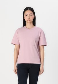 Różowa bawełniana koszulka z krótkim rękawem i kieszonką na piersi. Posiada okrągły dekolt i luźny krój, zestawiona z ciemnymi dżinsami.