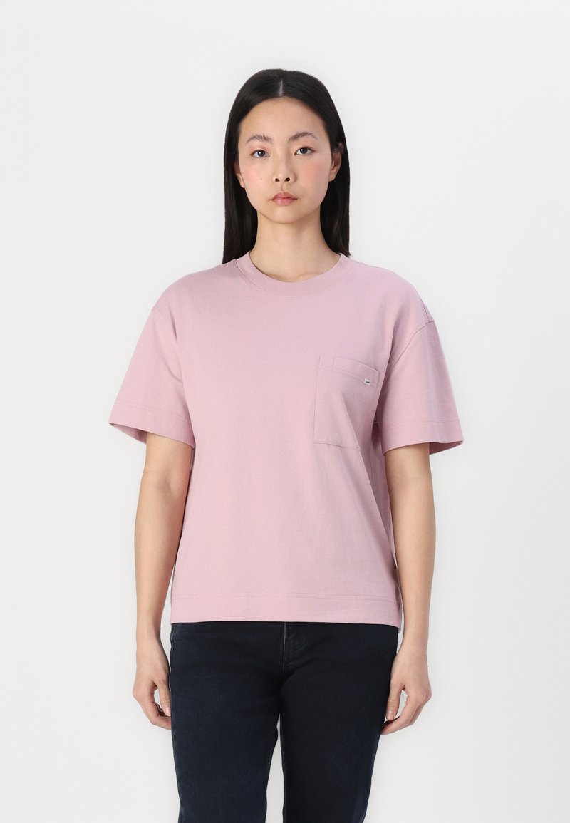 Różowa bawełniana koszulka z krótkim rękawem i kieszonką na piersi. Posiada okrągły dekolt i luźny krój, zestawiona z ciemnymi dżinsami.