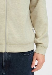 Light beige zip-up sweatshirt with a smooth texture, waistband, and cuffs, paired with dark blue jeans featuring a front pocket.