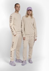 Il completo beige, composto da felpa e pantaloni, presenta motivi a zigzag marroni sulle maniche e sulle gambe; abbinato a sneaker viola chiaro e un cappellino coordinato.