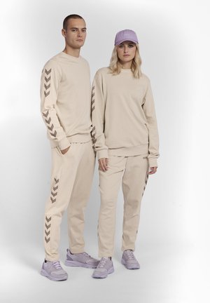 Il completo beige, composto da felpa e pantaloni, presenta motivi a zigzag marroni sulle maniche e sulle gambe; abbinato a sneaker viola chiaro e un cappellino coordinato.