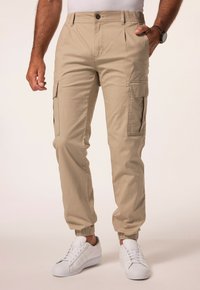 Beige cargo bukser med elastiske mansjetter, der har sidelommer og knaplukning. Lavet af tekstureret stof. Sættes sammen med hvide sneakers.