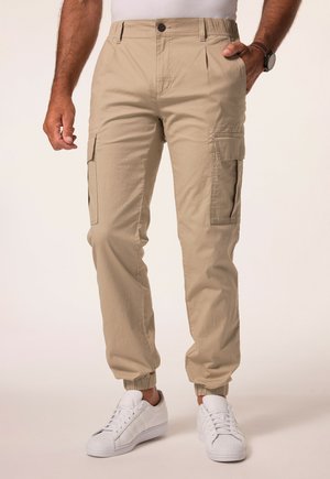 Pantaloni cargo beige con polsini elasticizzati, dotati di tasche laterali e chiusura a bottoni. Realizzati in tessuto testurizzato. Abbinati a sneakers bianche.