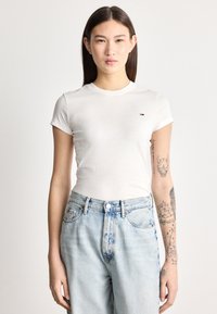 Jauna sieviete ar gariem tumšiem matiem, valkājot baltu pieguļošu T-kreklu un gaiši zilas džinsas, izdaiļojot ar tetovējumiem labo apakšdelmu.