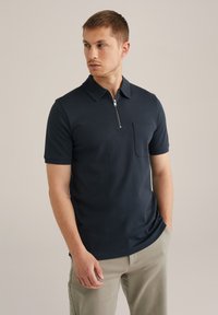 Polo de manga corta en color navy con cuello de media cremallera, bolsillo en el pecho y textura de tela suave, combinado con pantalones claros.