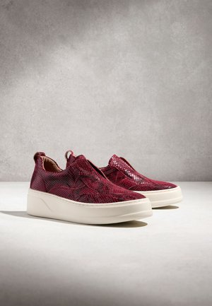 Slip-on tenisky s bordovou hadou textúrou a hrubými bielymi podrážkami na svetlom povrchu na sivom pozadí.