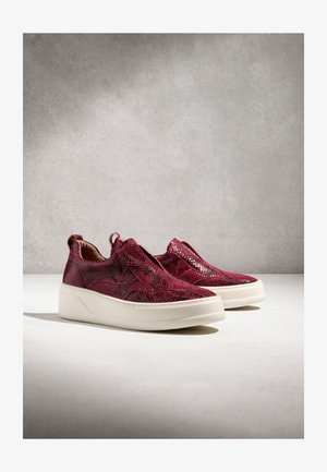 Slip-on tenisky s bordovou hadou textúrou a hrubými bielymi podrážkami na svetlom povrchu na sivom pozadí.