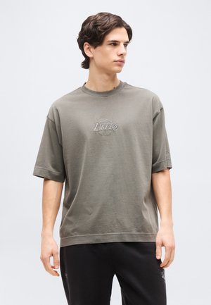 Mlad fant s temnimi lasmi, oblečen v preveliko sivo majico Los Angeles Lakers in črne hlače, gleda v stran na enostavni ozadju.