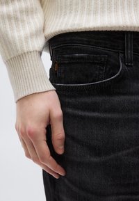 Fekete farmernadrág texturált felülettel, oldalzsebbel és egy kis narancssárga "BOSS" címkével, amely részben egy krémszínű bordázott pulóver által van takarva.