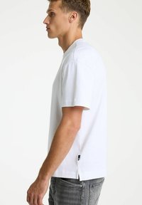 Camiseta de algodón blanco con un corte relajado, mangas cortas, aberturas laterales en el dobladillo y un detalle de etiqueta negra cerca de la parte inferior.